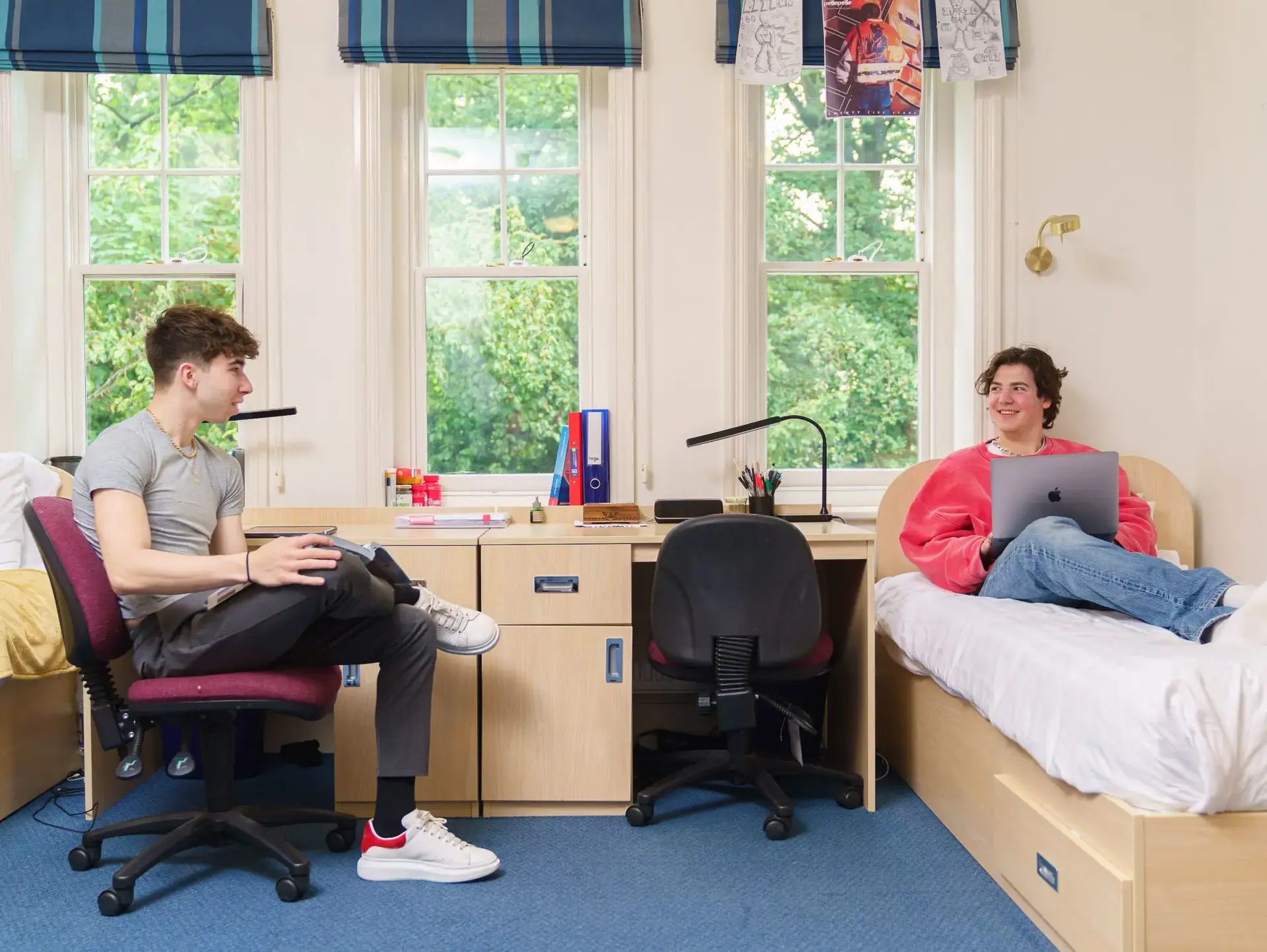 Two students sitting in their accommodation room having a conversation. One students sitting on the bed with a laptop and the other on a chair