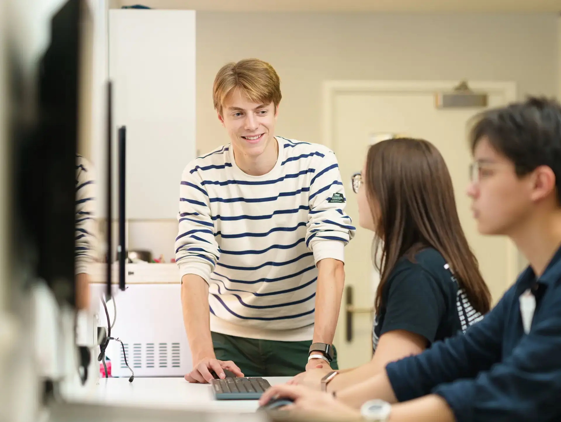 Students working at desk, with one other student standing and leaning over to chat