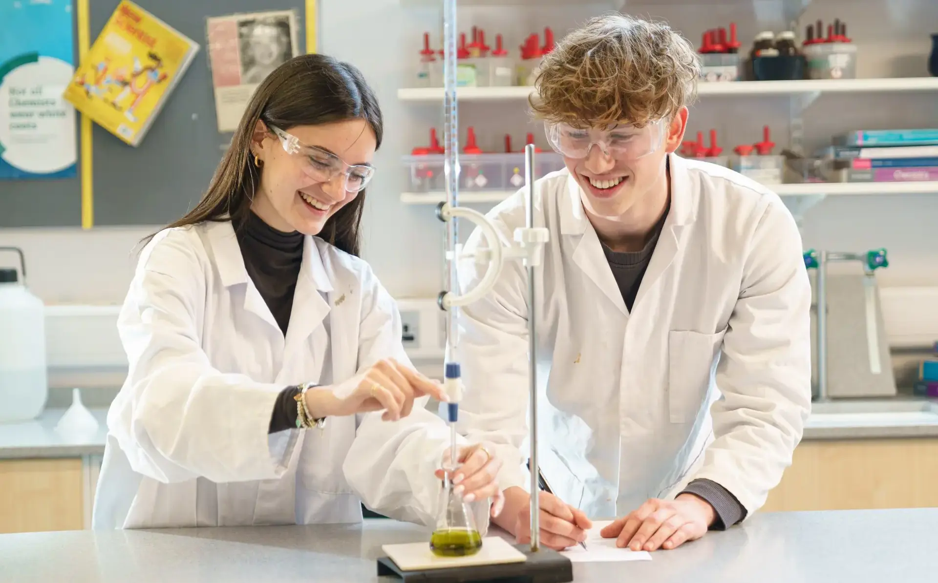 Two students wearing protective laboratory gear working together at table