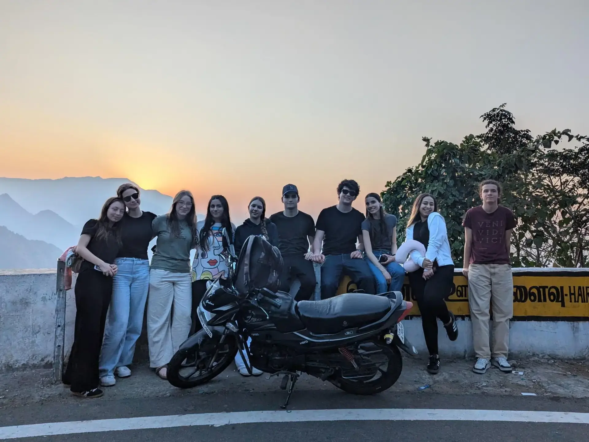 Students posing for group photo behind motorbike, with the sun setting in the background