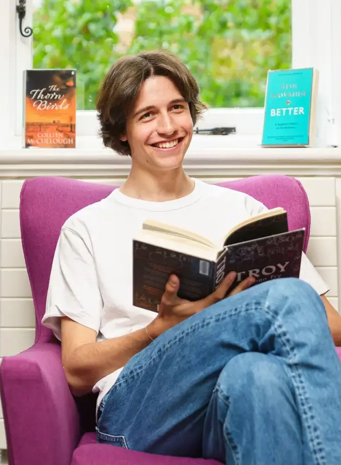 Male student sat in chair in library holding a book