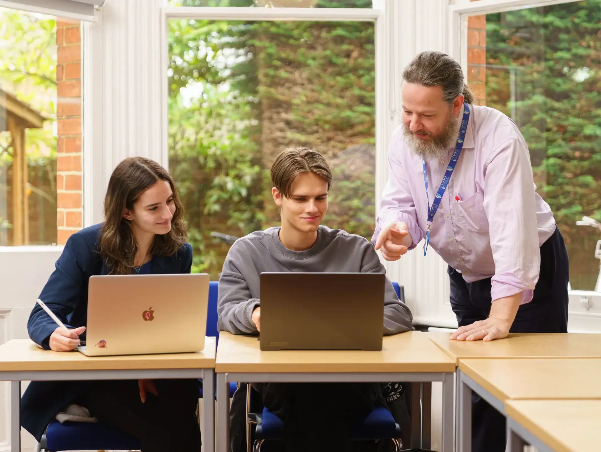 Teacher leaning over two students sitting at desk, discussing work on laptop