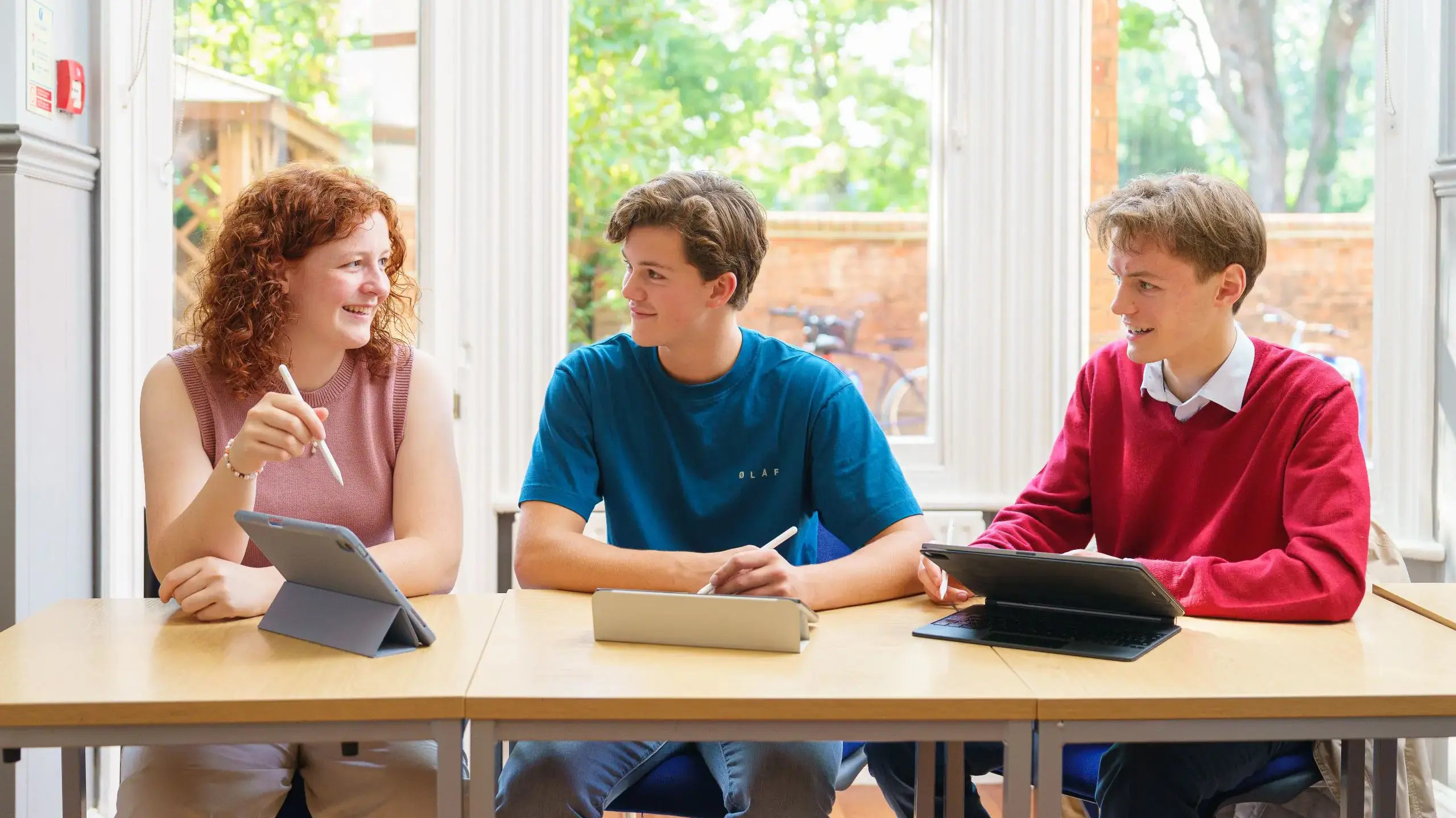 Three students working on tablets in front of large window